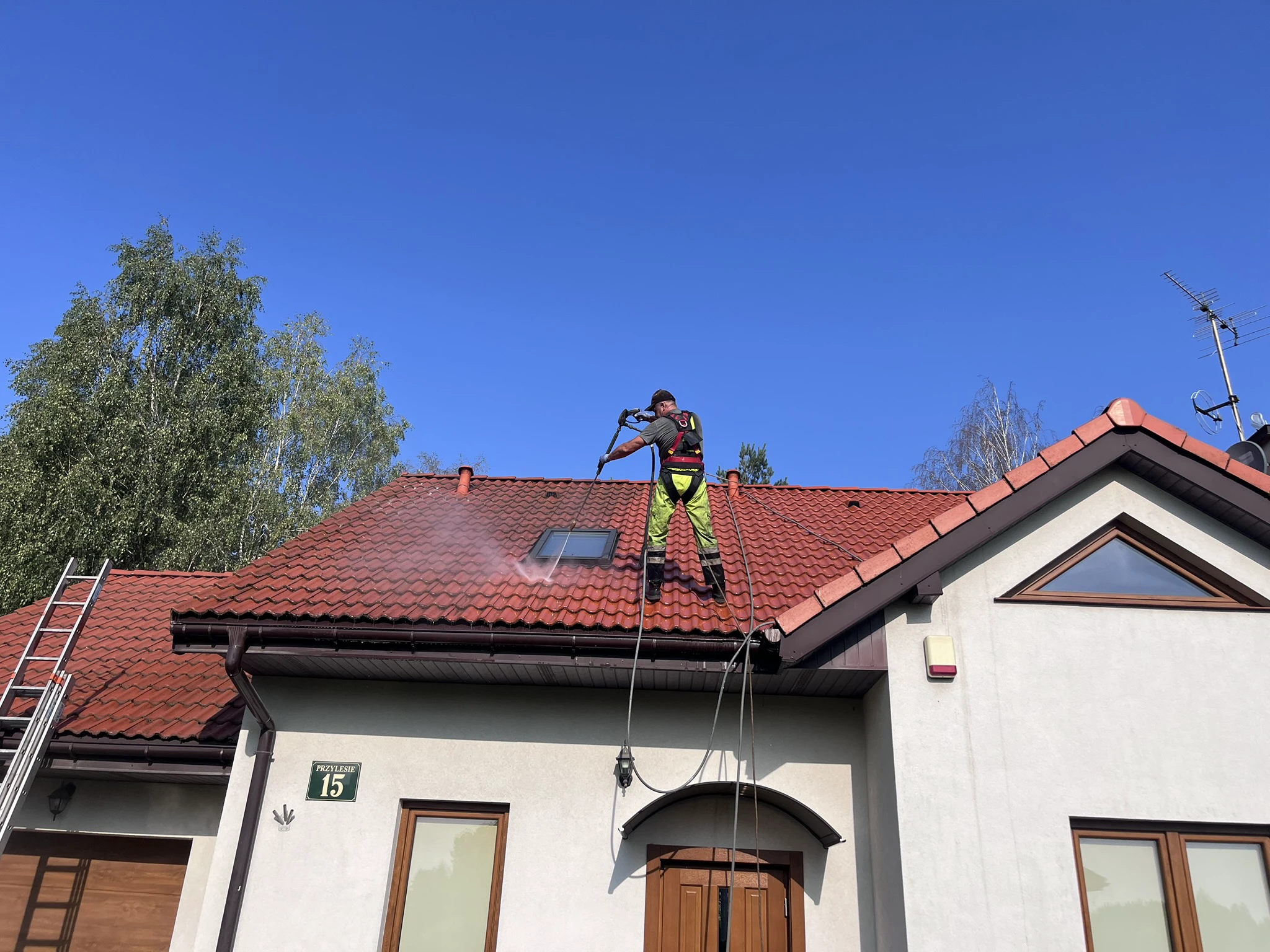 Pracownik na drabinie czyści dachówkę ciśnieniowo wodą na dachu budynku mieszkalnego – mycie dachu