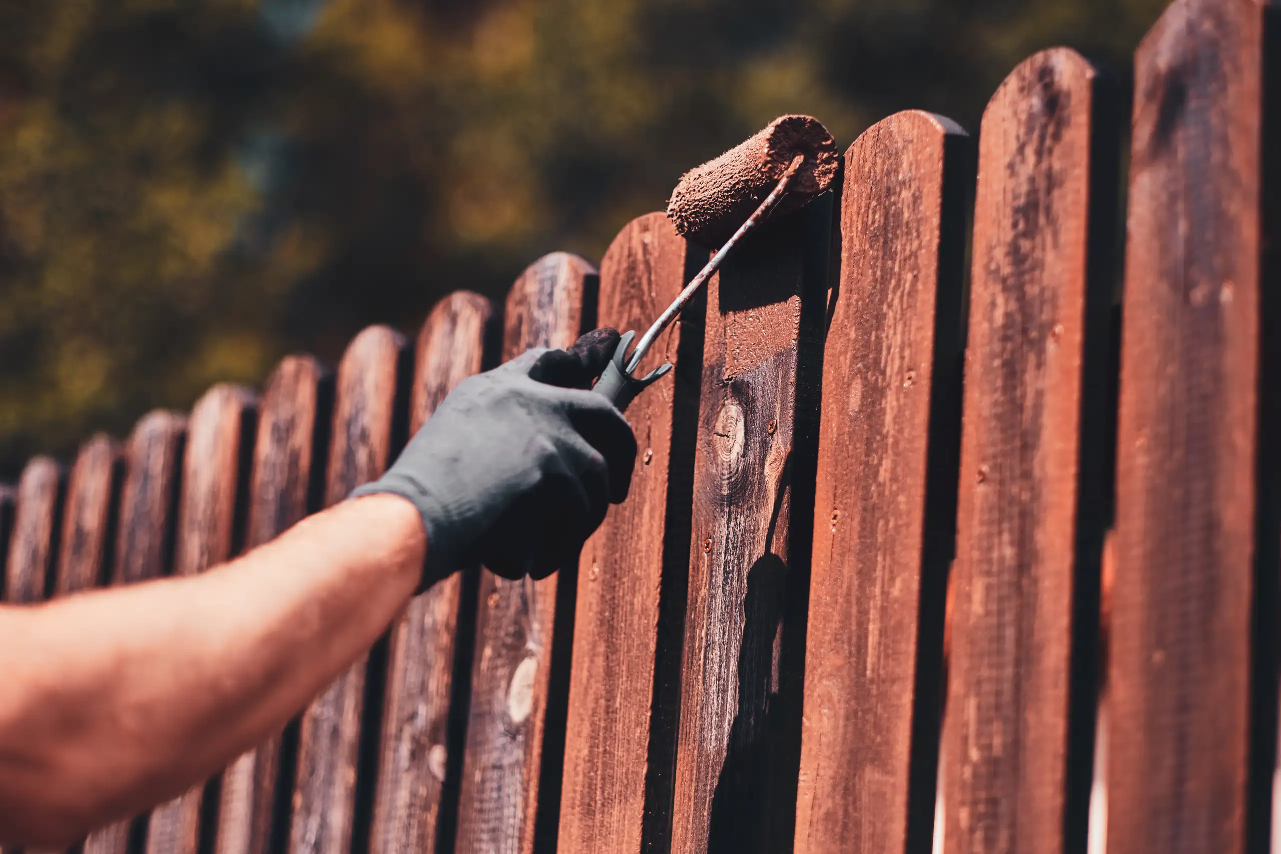 Malowanie ogrodzeń – pracownik w rękawicy nakłada brązową farbę wałkiem na drewniane przęsło ogrodzenia