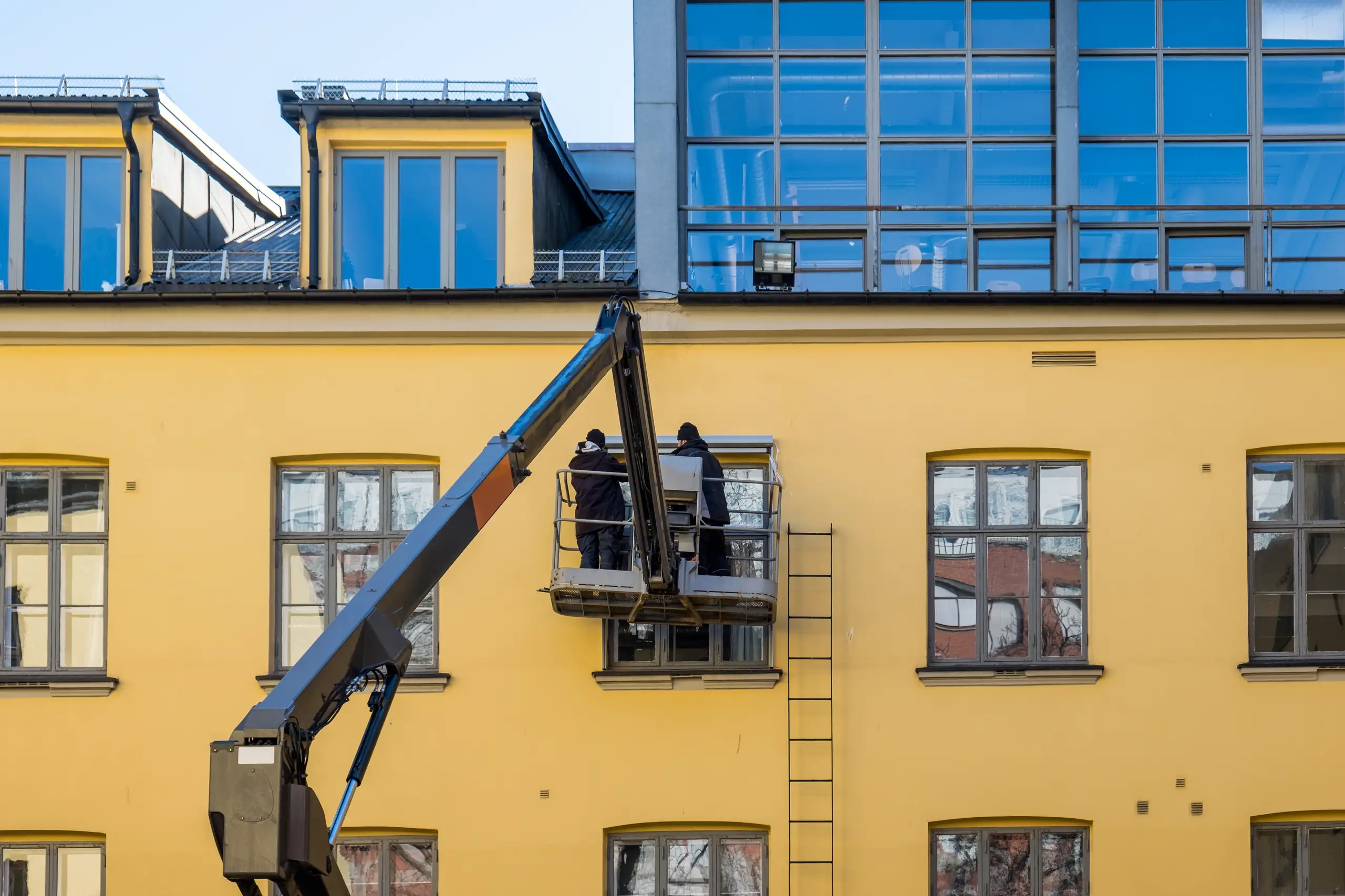 Dwie osoby z podnośnikiem koszowym wykonują Mycie elewacji żółtej ściany budynku